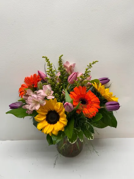 Bright spring arrangement — sunflowers, orange gerbera daisies, purple tulips and pink alstroemeria in a glass vase