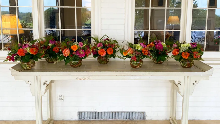 Seven coordinated orange-and-pink centerpieces lined along a cream console table on a clubhouse porch with tall windows