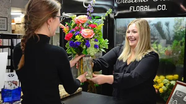 A smiling staff member hands a pink and purple arrangement across the black counter to a customer, vintage Crescent Street Floral Co. signage above the cooler behind them