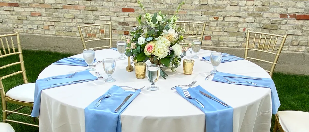 Round wedding table set with white linens, periwinkle napkins and gold chiavari chairs around a blush centerpiece, against a cream brick wall